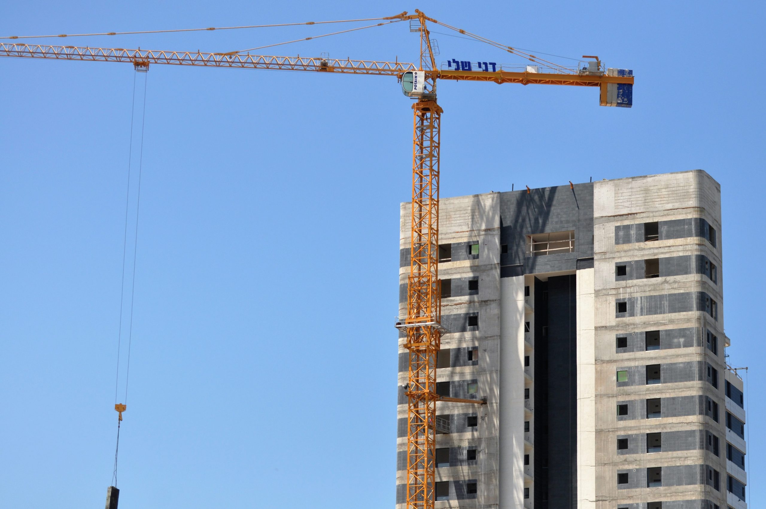 בניין בתהליך בנייה עשוי בטון חשוף לצד מנוף עם שלט של חברת דני שלי מהנדסים, פרויקטים הנדסיים, ביצוע קבלנות בניין, פיתוח תשתיות, איכות בנייה גבוהה, חדשנות בתחום הבנייה.